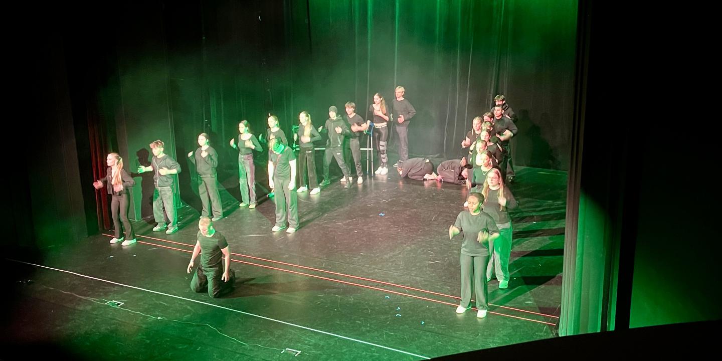 Aufführung 8: Die Freikugeln  – Die Gruppe steht auf der Bühne in einer V-Formation. Es leuchtet grünes Licht. Ein einzelner Darsteller kniet am Bühnenrand.